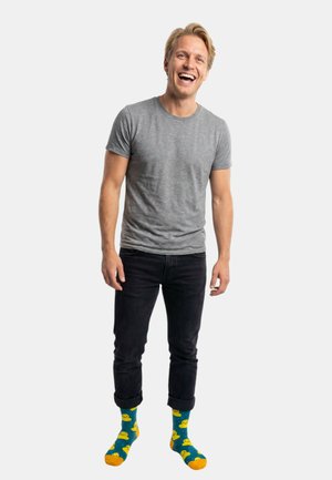 Lächelnder Mann, der ein graues T-Shirt, schwarze hochgekrempelte Hose und blaue Socken mit gelben Gummienten-Mustern trägt, vor einem weißen Hintergrund steht.