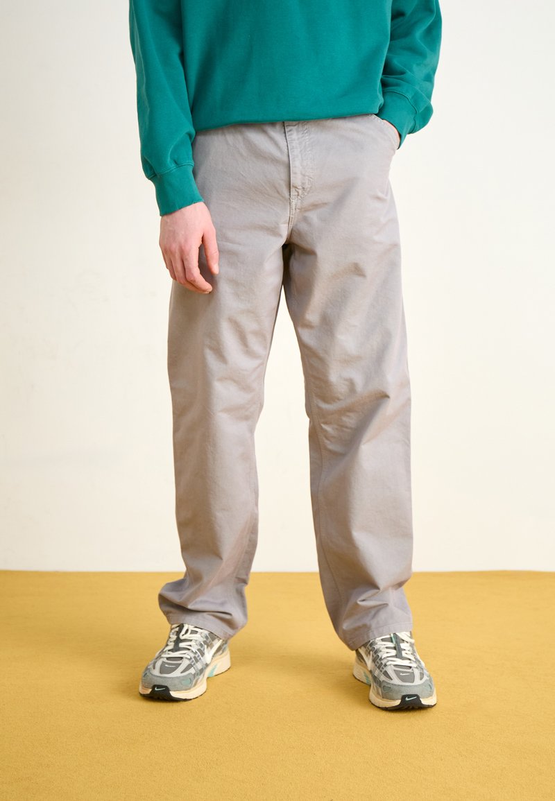 Lichtgrijze, losse broek van katoen met een gladde textuur, gecombineerd met multikleuren sport schoenen en een groene top met lange mouwen.