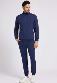 Marineblauer Trainingsanzug mit einer Jacke mit Reißverschluss und passenden Hosen. Die Jacke hat einen hohen Kragen und seitliche Taschen, die schwarze Akzente aufweisen. Weiße Sneaker runden den Look ab.