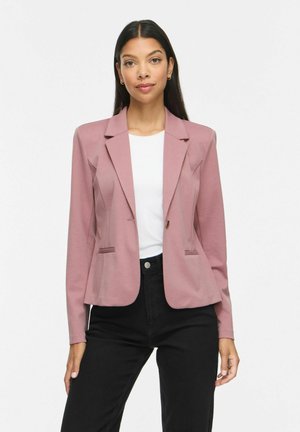 Kvinde iført en tætsiddende støvet lyserød blazer over en hvid top og sorte højtaljede bukser, stående mod en ensartet lys baggrund.