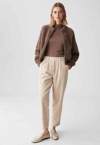 Vrouw in een bruine fuzzy jas over een bruine coltrui, beige geplooide corduroybroek en beige instappers, staand met handen in haar zakken.
