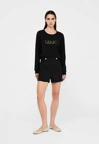 Czarna bluza z brokatowym logo, zestawiona z czarnymi szortami z ozdobnymi guzikami. Modelka stoi z neutralnym wyrazem twarzy, ubrana w beżowe baleriny.