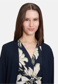 Marineblauwe cardigan over een bloemenpatroon jurk met een geknoopte taille, met grote witte bloemen en groene bladeren tegen een donkere achtergrond.