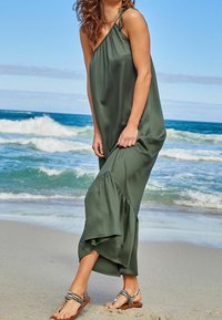 Robe maxi sans manches vert olive avec un ourlet à volants. Fabriquée en tissu lisse, elle présente un détail de lien sur le côté. Associée à des sandales rayées.