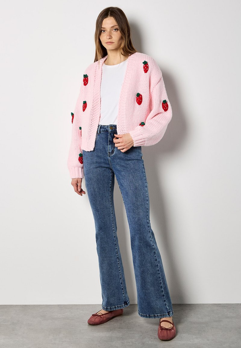 Cardigan rose tricoté avec des appliqués de fraises, assorti à un jean évasé bleu et des ballerines rouges. Tenue décontractée avec une coupe relaxée.