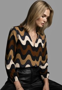 Mehrfarbige Bluse mit einem wellenförmigen Muster in Braun-, Schwarz- und Cremetönen, mit V-Ausschnitt und elastischen Bündchen, kombiniert mit schwarzen Lederhosen.