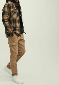 Persona in pantaloni cargo beige, sneakers bianchi, felpa nera e giacca a quadri marrone che si trova contro uno sfondo semplice.