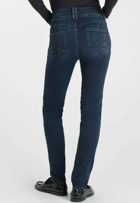 Personne portant un jean slim-fit bleu marine et des mocassins noirs, vue de dos contre un fond clair uniforme.