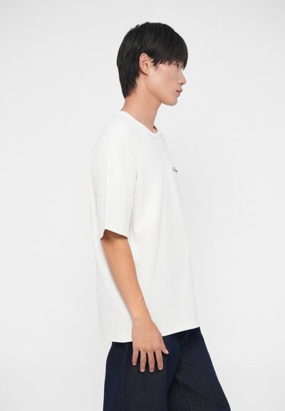 Camiseta blanca de algodón con un corte holgado y mangas cortas. Presenta un cuello redondo y un pequeño logo en el pecho. Se lleva con jeans oscuros.