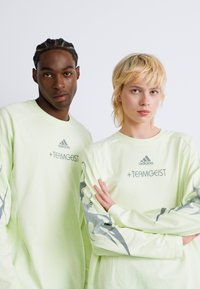 Dva mlada odrasla lica nose svijetlozelene Adidas +TEAMGEIST dukserice i stoje protiv jednostavne bijele pozadine.