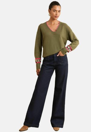 Donna che indossa un maglione verde oliva con scollatura a V e maniche con stampe rosa e leopardate, jeans neri a gamba larga e scarpe a punta color cammello.