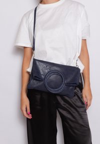 Marineblauwe leren handtas met een cirkelvormige accent, geplooid ontwerp en een schouderriem, met een gladde textuur en glanzende afwerking.