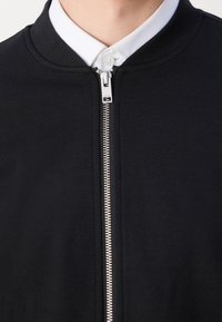 Gros plan sur une personne portant une veste zippée noire par-dessus une chemise blanche à col, avec un accent sur la partie supérieure de la poitrine et la fermeture éclair.