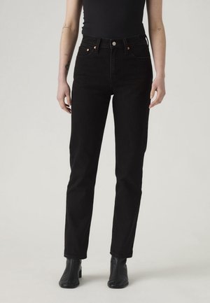 Osoba ubrana w czarne dżinsy z wysokim stanem, czarny bezrękawnik i czarne botki, stojąca na tle jednolitego jasnego tła.