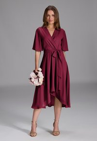 Robe enveloppante bordeaux à manches courtes, taille cintrée et ourlet asymétrique. Associée à des sandales dorées à lanières et à un bouquet floral.