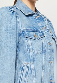 Světle modrá džínová bunda s knoflíkovým zapínáním, dvěma kapsami na prsou a detailem na rukávech s nařasením. Má klasický límec a vybledlou texturu.