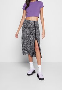 Femme portant un crop top violet, une jupe midi à motifs noir et blanc avec une fente latérale, des chaussettes Nike blanches et des mocassins chunky lilas, se tenant sur un sol gris.
