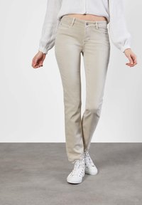 Jeans beige de pierna recta con cinco bolsillos, cremallera y cierre de botón. Combinados con zapatillas altas blancas, en un fondo neutro.