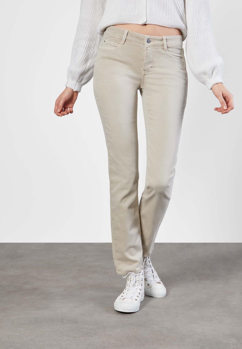 Jeans beige de pierna recta con cinco bolsillos, cremallera y cierre de botón. Combinados con zapatillas altas blancas, en un fondo neutro.