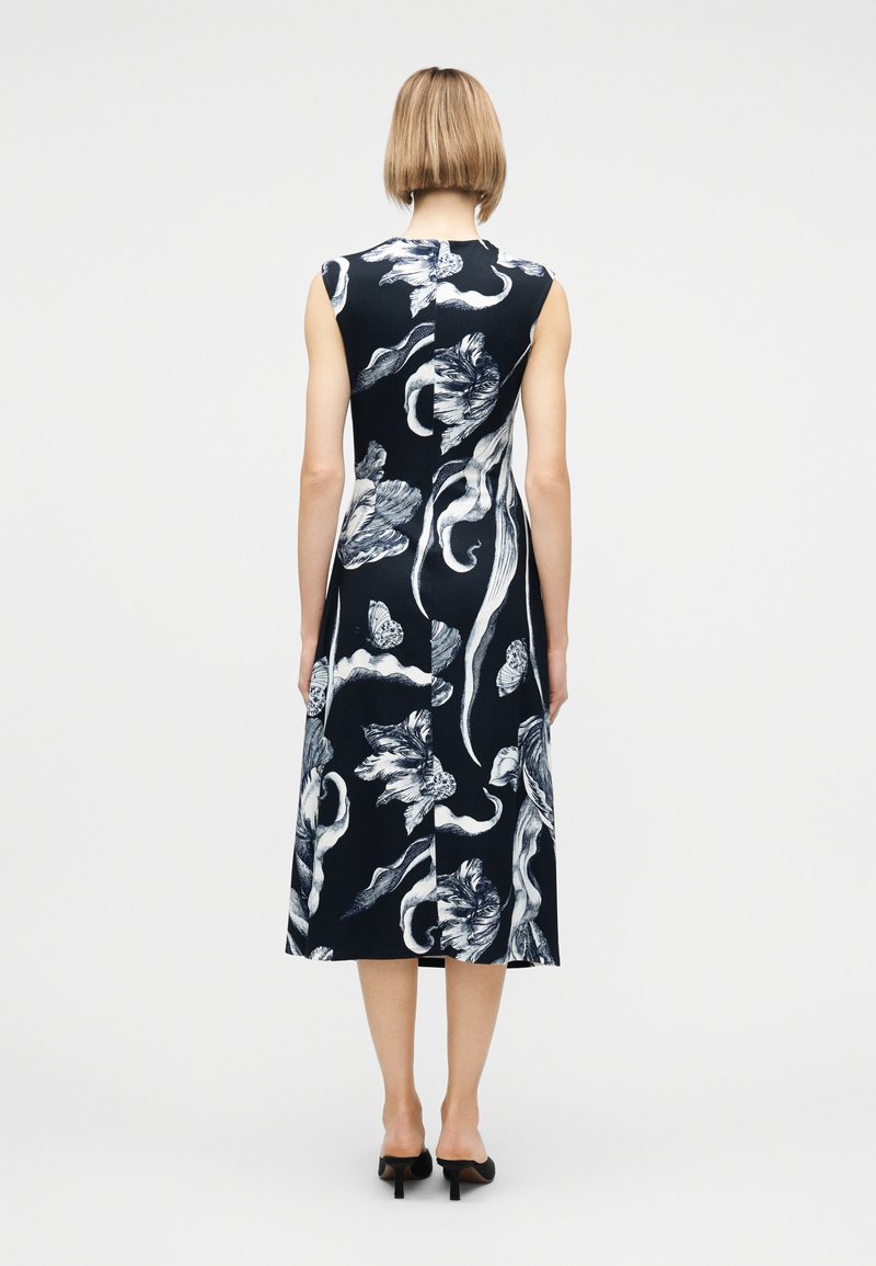 Woman with short blonde hair wearing a sleeveless black midi dress with white floral and abstract patterns, standing with back facing forward.