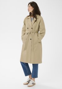 Femme portant un trench-coat beige avec ceinture, jean bleu coupé, baskets noir et blanc, regardant sur le côté sur un fond uni.