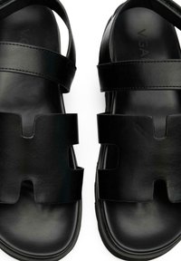 Black leather sandals featuring wide straps with cut-out shapes, smooth texture, and a flat sole design.