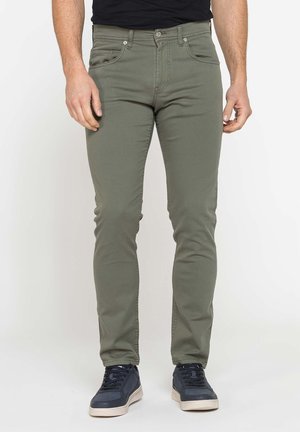 Uomo con pantaloni slim verde oliva e scarpe da ginnastica nere in piedi su uno sfondo chiaro e uniforme.