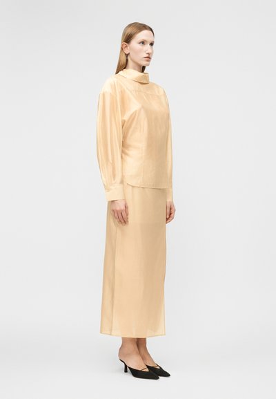 Γυναίκα που στέκεται φορώντας μπεζ μπλούζα με μακριά μανίκια και ψηλό λαιμό και φούστα μήκους αστραγάλου στο ίδιο χρώμα, με μαύρες μυτερές γόβες, στραμμένη προς τα δεξιά.