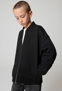 Giacca bomber nera con zip, caratterizzata da un collo alto, vestibilità oversize e un discreto logo ricamato sul petto. Texture liscia con polsini elasticizzati.