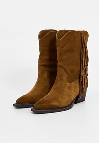 Brązowe zamszowe kowbojskie buty z wydłużonym noskiem, na blokowych obcasach, z frędzlami po zewnętrznych stronach, przedstawione na białym tle.