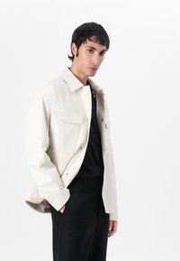 Lichtbeige ritjack met kraag, twee borstzakken en een losse pasvorm, gecombineerd met zwarte broek met streepjes.