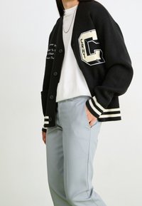 Zwarte cardigan met witte geribbelde manchetten en zoom, voorzien van een grote "C" patch en geborduurde tekst, gecombineerd met lichtgrijze broeken.