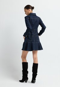 Robe en denim avec un corsage ajusté, une jupe évasée et deux poches arrière. Dispose d'un col, de manches longues et d'une taille ceinturée. Bottes noires jusqu'aux genoux.