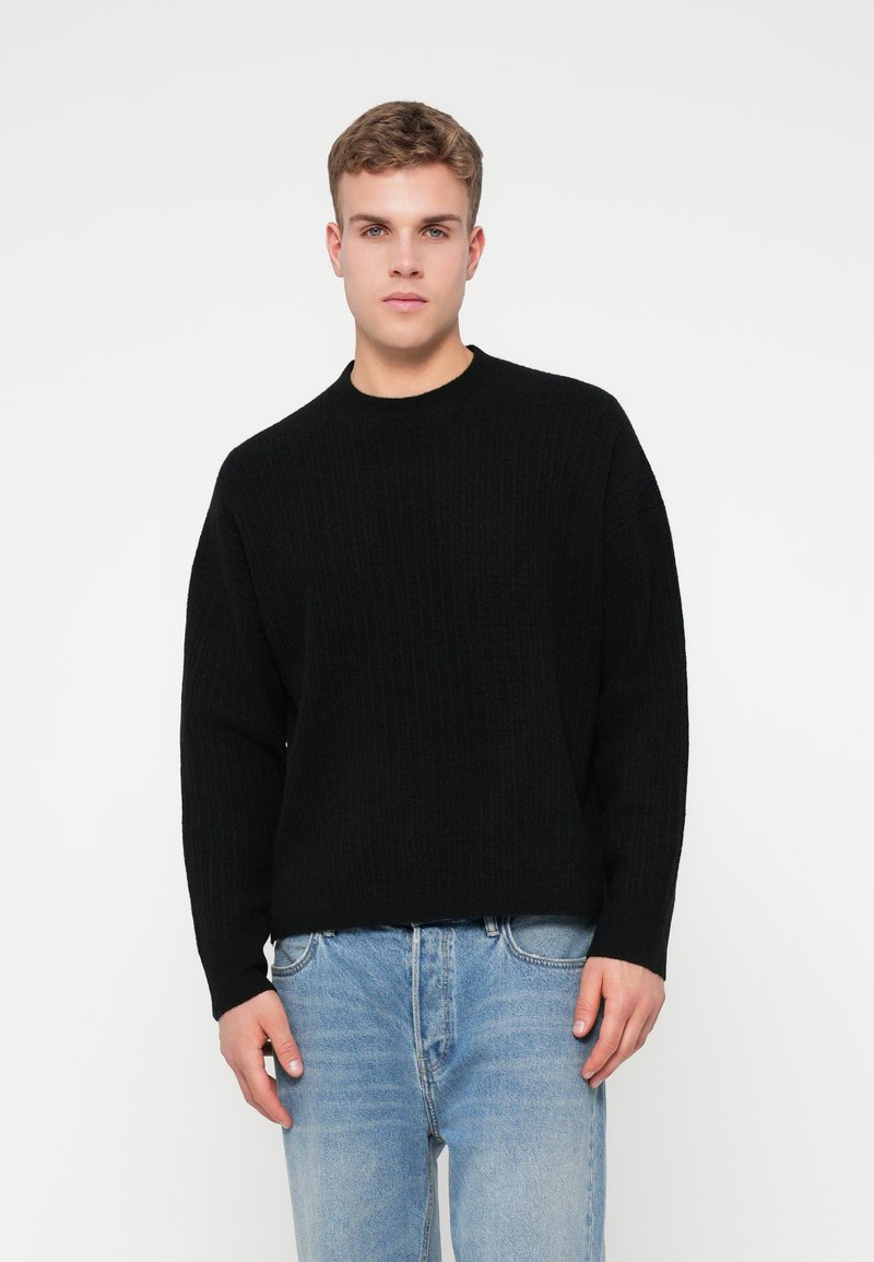 Suéter negro de punto acanalado con cuello redondo, mangas largas y corte relajado, combinado con jeans de mezclilla azul claro.