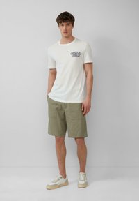 T-shirt en coton blanc avec un motif graphique bleu sur la poitrine, associé à un short vert olive et des baskets claires.