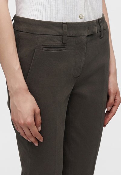 Personne portant un pantalon marron foncé sur mesure et un haut blanc côtelé, les mains reposant le long du corps.