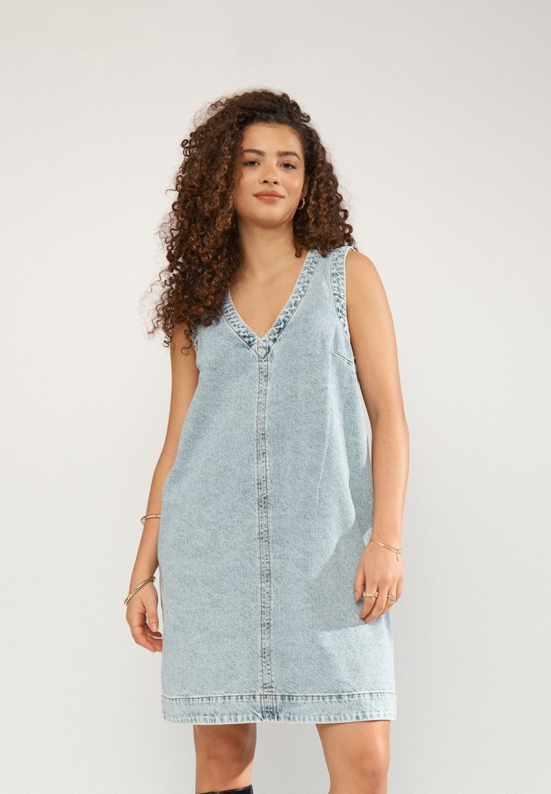 Jeune femme aux cheveux bouclés portant une robe en denim bleu clair sans manches, debout devant un fond uni.