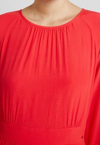 Rotes Kleid mit runder Ausschnitt, geraffter Textur an der Passe und einer enganliegenden Taille. Der Stoff wirkt weich und leicht.