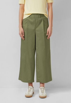 Person trägt olivgrüne Hosen mit weitem Bein, gelbes kurzärmeliges Oberteil und weiße Turnschuhe und steht vor einem schlichten Hintergrund.