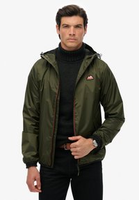 Olivgrön zip-jacka med huva, lättviktig nylon, elastiska ärmmuddar och röda accenter. Har en triangulär logotyp på bröstet.