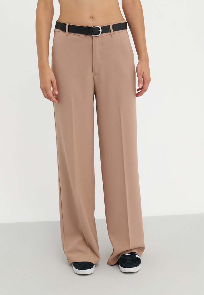 Pantalones de pierna ancha en tela de color beige claro, que presentan un cinturón negro con una hebilla de metal. Textura suave, diseño ajustado y pliegues frontales visibles.