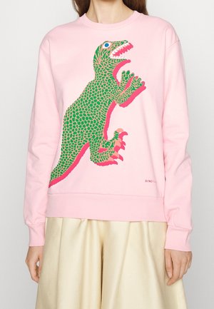 Personne portant un sweat-shirt rose clair avec un grand dessin de dinosaure de style cartoon en vert et rose à l'avant, associé à un pantalon beige.
