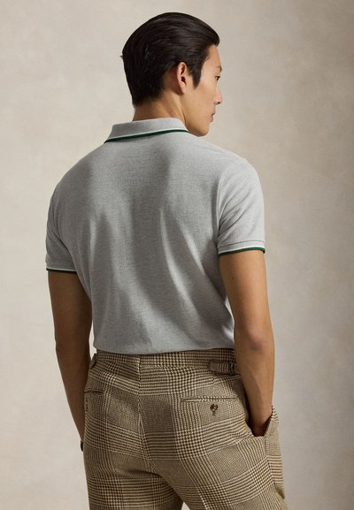 Homme aux cheveux foncés se tenant de dos, portant un polo gris clair avec une bordure verte et un pantalon beige à carreaux, les mains dans les poches.