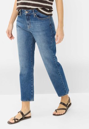 Person iført blå jeans med lige ben, brunt og hvidt stribet top og mørkebrune flade sandaler med guldlås, stående på hvidt gulv.