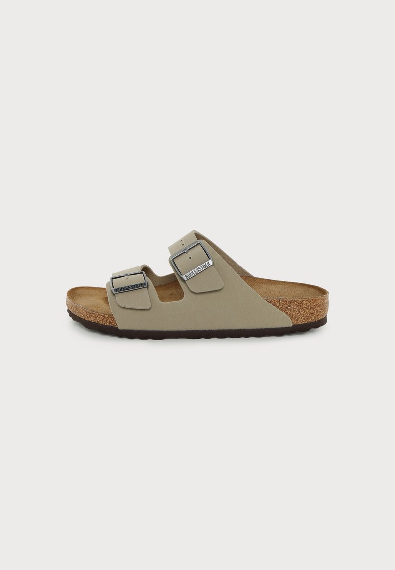 Μπεζ σανδάλι Birkenstock με δύο ρυθμιζόμενους ιμάντες με αγκράφες, φελλό πάτο και σκούρη καφέ υφή σόλας σε μονόχρωμο φόντο.