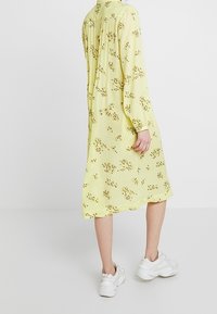 Donna che indossa un abito giallo largo a maniche lunghe con piccoli motivi floreali e sneakers bianche chunky, in piedi contro uno sfondo chiaro e semplice.