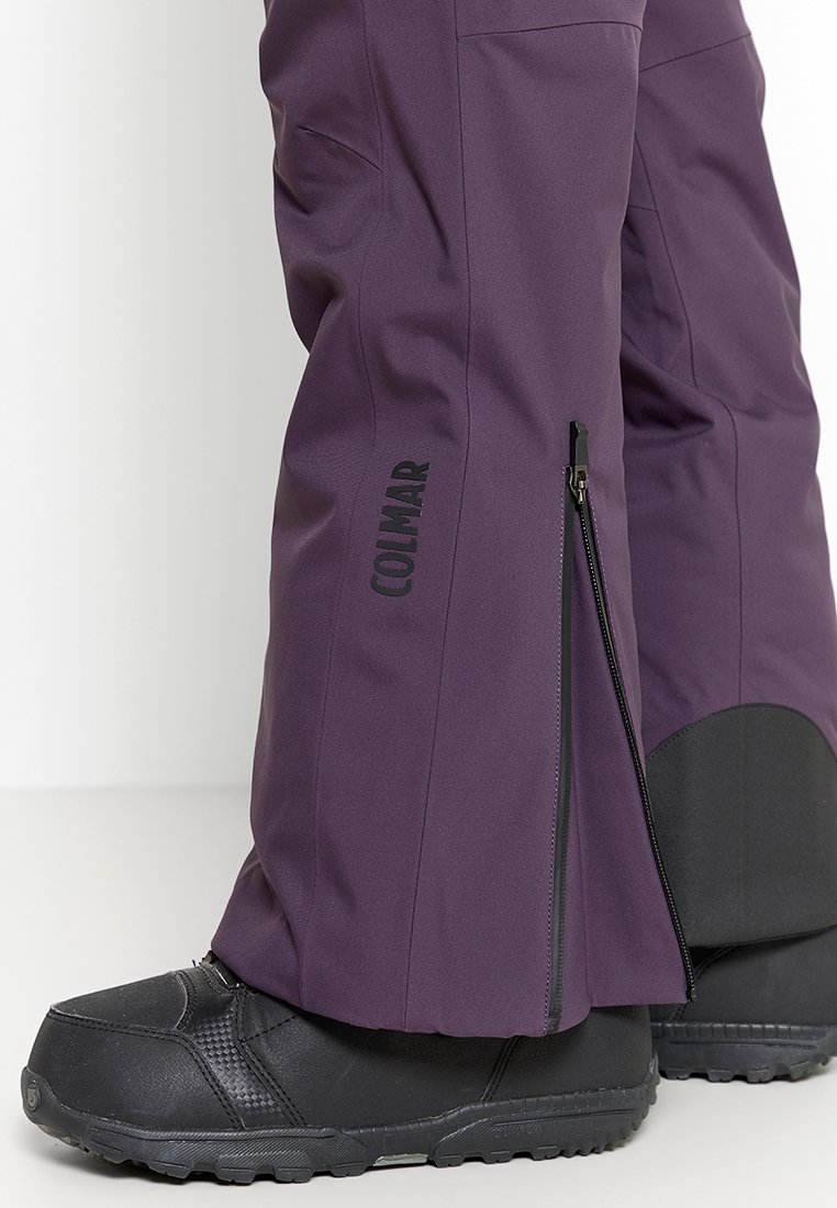 Lila Skihosen mit seitlichem Reißverschluss über schwarzen Schneestiefeln, die eine strukturierte Sohle und das "COLMAR"-Logo am Hosenbein zeigen.