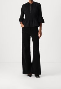 Schwarze plissierte Bluse mit hohem Kragen und Vorderreißverschluss, kombiniert mit schwarzen weit geschnittenen Hosen und schwarzen Pumps mit spitzem Zeh. Glatte Textur über die gesamte Oberfläche.
