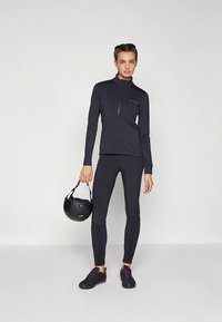 Długi rękaw, podpinana ciemnogranatowa sportowa bluza w połączeniu z dopasowanymi legginsami. Czarny kask trzymany w jednej ręce, buty z fioletowymi akcentami.