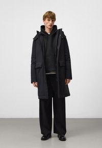 Czarna kurtka puchowa z kapturem, dwoma dużymi przednimi kieszeniami i zapięciem na zamek. Noszona na ciemnej bluzie z kapturem i luźnych czarnych spodniach.